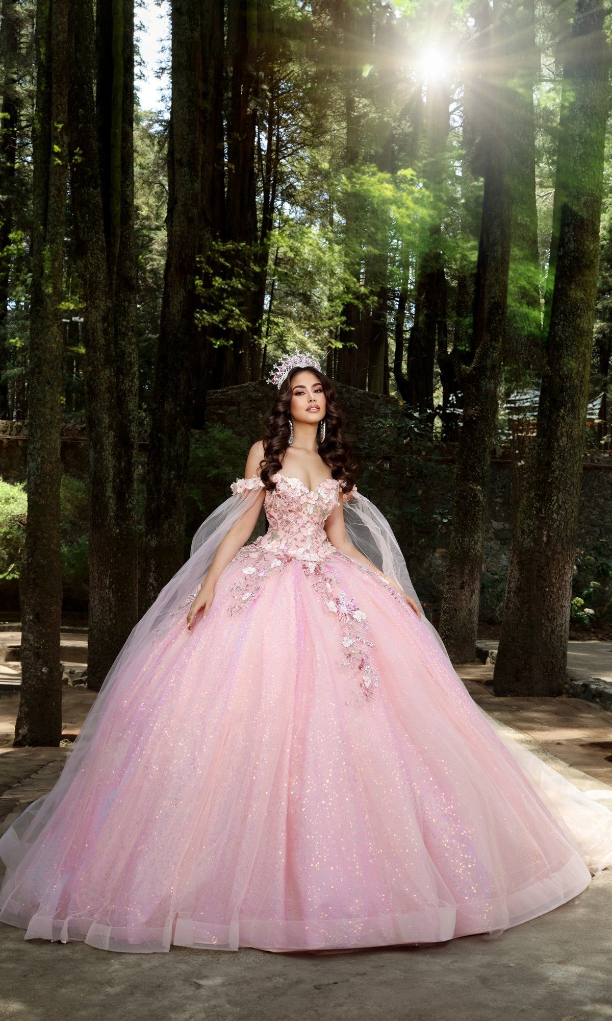 A woman wearing the Quinceanera Dress Q114 by Nina Canacci in blush, featuring floral appliques and a glitter tulle skirt, stands in a sunlit forest among tall trees.